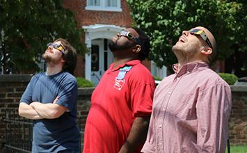 Three men wearing solar eclipse glasses looking up at the sun